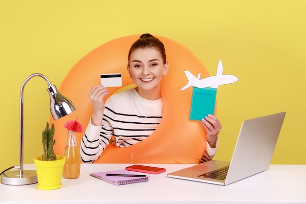 Happy woman with rubber ring showing credit card and passport booking tickets online on laptop