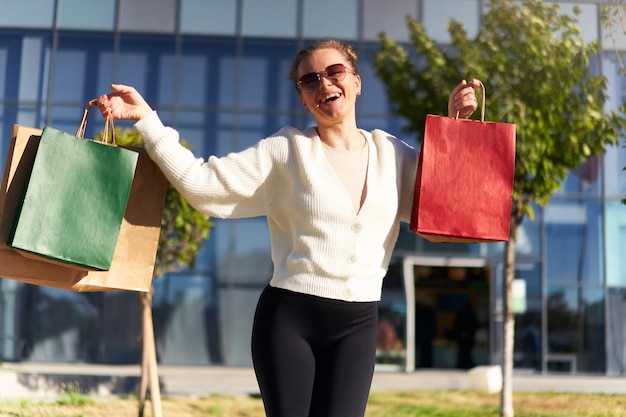 Happy woman with shopping bags walking in city from mall Excited girl enjoys purchases on black friday sale Female posing on camera with full paper bags Consumerism concept