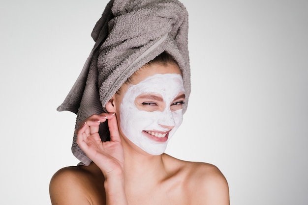 Happy woman with a towel on her head applied a mask to clean the face skin on a gray background