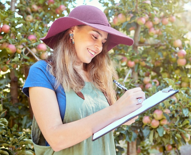 Happy woman writing notes on clipboard while working on sustainable apple orchard farm on sunny day Cheerful farmer reading checklist while planning task for harvest season of fresh organic fruit