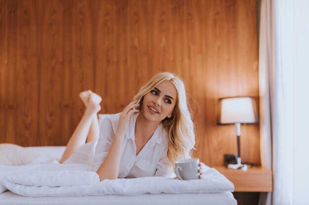 Happy womanl using a mobile phone while lying on the bed and drink morning coffee at home