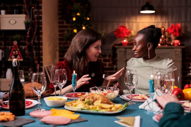 Happy women chatting at christmas home party, cheerful friends talking at festive dinner table, eating traditional xmas food. Winter holiday celebration in beautiful decorated place