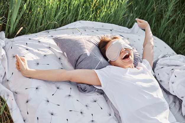 Happy yawning female wearing sleeping fold and white casual t-shirt stretching arms when waking up in morning in bed in green meadow, relaxing and sleeping outdoor in field.