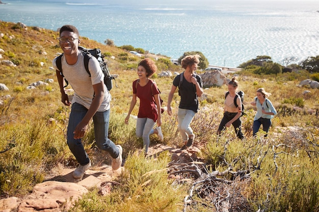 Happy young adult friends hiking single file uphill on a path by the coast full length