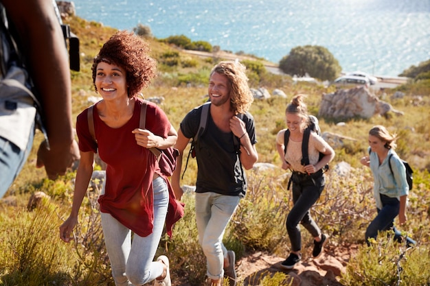 Happy young adult friends hiking single file uphill on a path by the coast full length