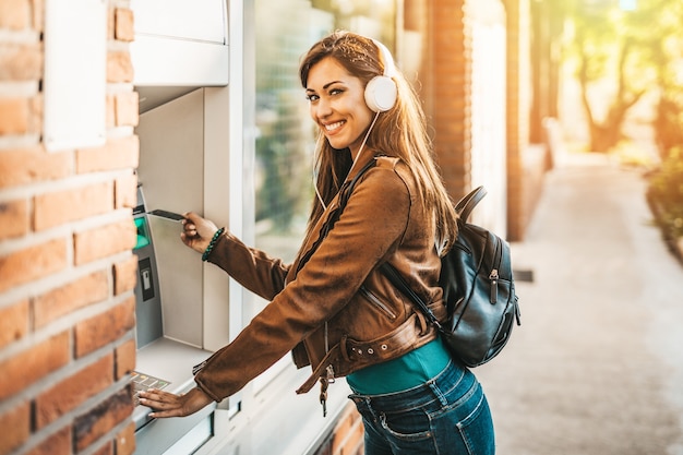 Happy young adult woman with headphones on her head standing in front of atm machine, smiling and holding credit or debit card.