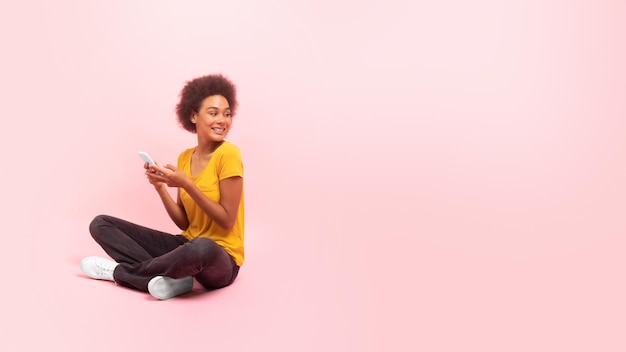 Happy young african american curly lady sit on floor look back at empty space isolated on pink background