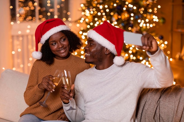 Happy young african american family in Santa hats cheers glasses with champagne make selfie on phone