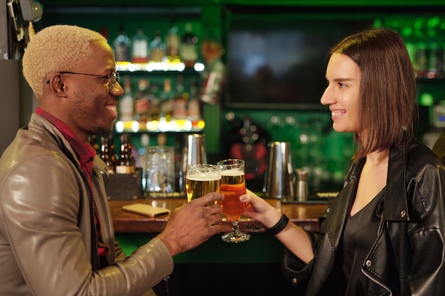 Happy young African man in smart casualwear looking at his girlfriend with smile while clinking with glasses of beer in front of camera in bar