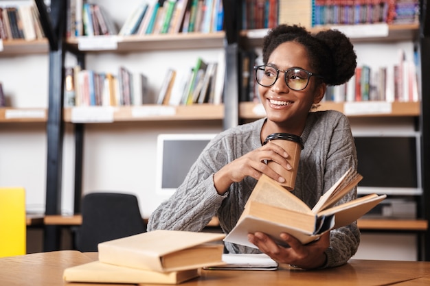 Happy young african student girl studying at the library, reading a book