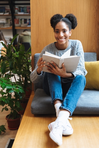 Happy young african student girl studying at the library, reading a book