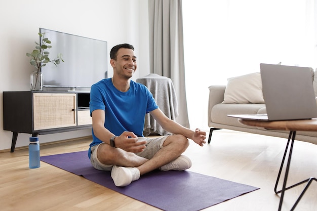 Happy young arab man in sportswear practicing yoga in lotus position looking at laptop screen and smiling