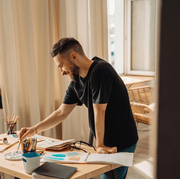 A happy young artist smiling makes a sketch with a brush in the studio A creative person is standing over a desktop with a brush in his handStudio lifestyle process of creating a work of art