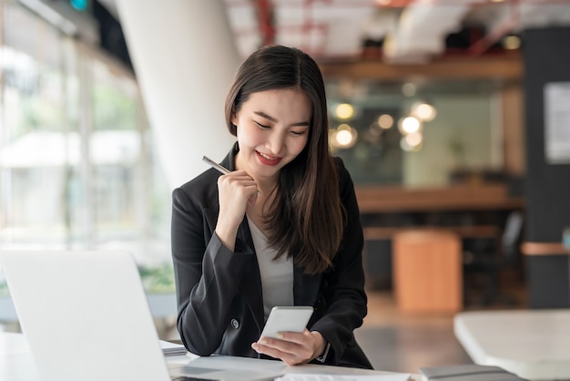 Happy young Asian businesswoman holding a pen using a smartphone at the modern office.