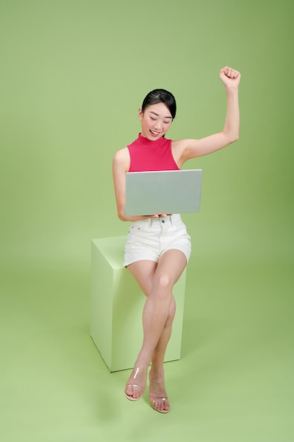 Happy young Asian businesswoman using laptop and sitting on podium isolated on green background