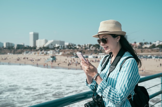 Happy young asian chinese girl backpacker looking mobile phone screen in beautiful summer sunny day near sea port. cheerful woman travel photographer holding cellphone and leaning on railing.