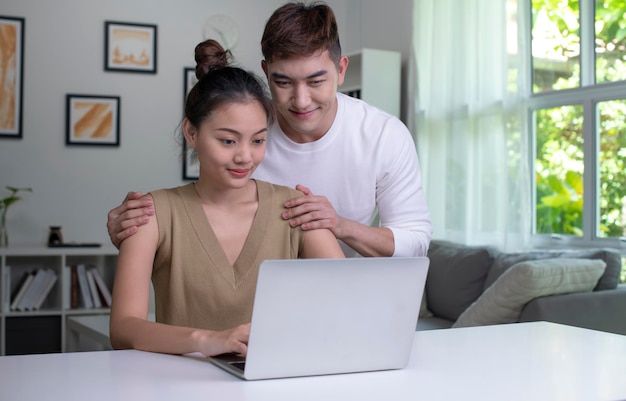 Happy young Asian couple communicating online on laptop Loving Woman and Man embracing watching movie webinar on laptop studying elearning together doing paperwork paying bills online in home office