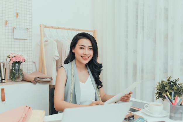 Happy young Asian designer in casualwear sitting by desk and making fashion sketches