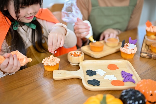 Happy young Asian girl enjoys decorating and making Halloween cupcakes with her mom