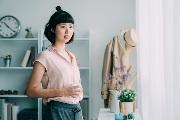 happy young asian japanese woman holding glass of water looking away at home. charming girl in short hair smiling watching city view by window in modern cozy apartment. lady enjoy morning indoors.