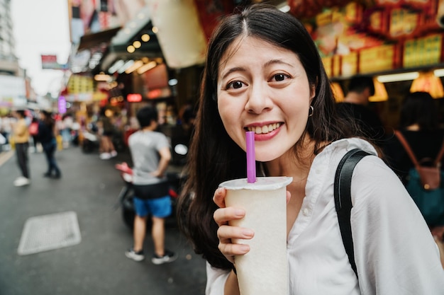 Happy young Asian korean woman traveler with bubble milk tea face camera smiling cheerful. beautiful lady tourist enjoy boba drink in disposable paper cup and straw on street outdoor market.