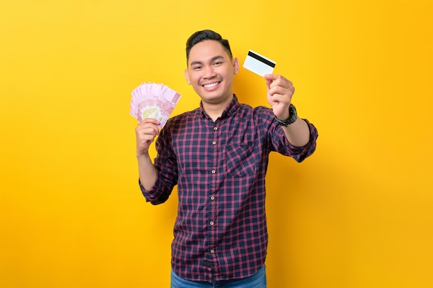 Happy young Asian man holding money banknotes and credit card isolated over yellow background Easy payment concept