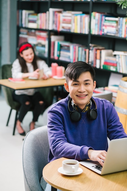 Happy young Asian man putting hands on the laptop keyboard and smiling while sitting in the Internet cafe with a cup of coffee