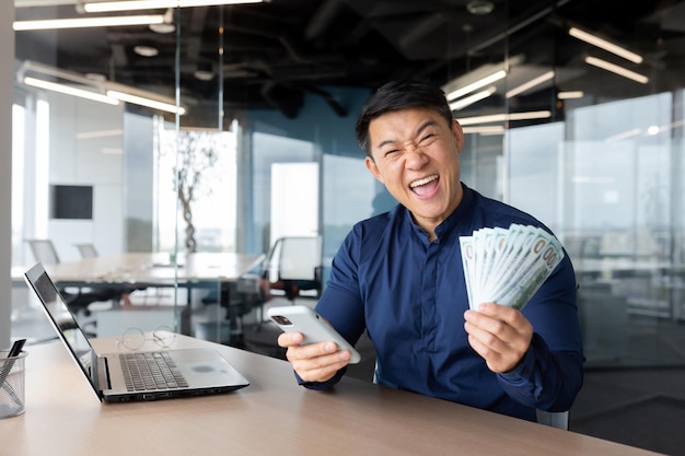 Happy young asian man sitting in office holding cash money and phone he looks at the camera shouts