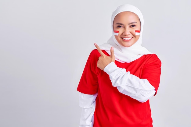 Happy young Asian muslim woman in red white tshirt pointing fingers aside at copy space isolated on white background Indonesian independence day on 17 august concept