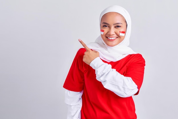 Happy young Asian muslim woman in red white tshirt pointing fingers aside at copy space isolated on white background Indonesian independence day on 17 august concept