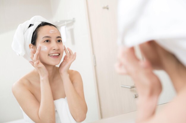 Happy young Asian woman applying face lotions while wearing a towel and touching her face in bathroom