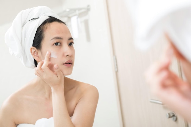 Happy young Asian woman applying face lotions while wearing a towel and touching her face in bathroom