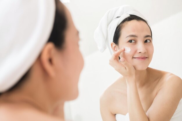 Happy young Asian woman applying face lotions while wearing a towel and touching her face in bathroom