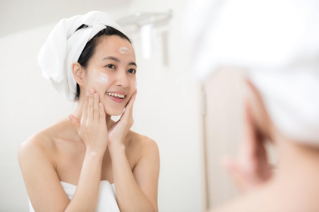 Happy young Asian woman applying face lotions while wearing a towel and touching her face in bathroom