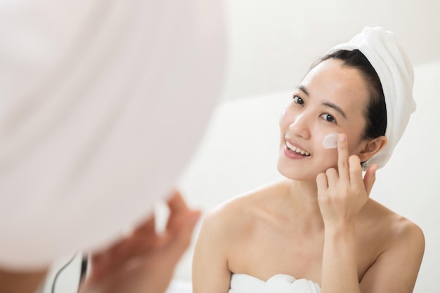 Happy young Asian woman applying face lotions while wearing a towel and touching her face in bathroom