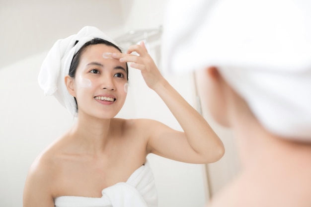 Happy young Asian woman applying face lotions while wearing a towel and touching her face in bathroom