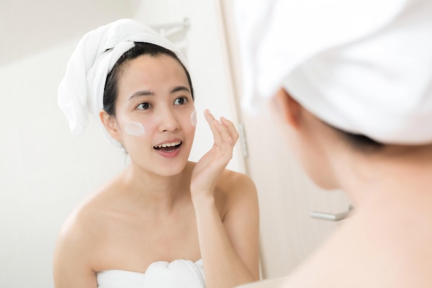 Happy young Asian woman applying face lotions while wearing a towel and touching her face in bathroom