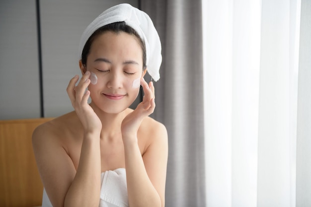 Happy young Asian woman applying face lotions while wearing a towel and touching her face Daily makeup and skincare