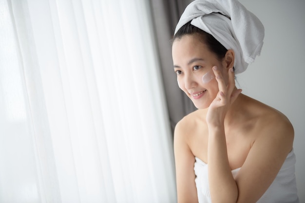 Happy young Asian woman applying face lotions while wearing a towel and touching her face Daily makeup and skincare