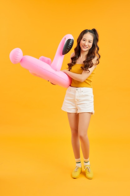 Happy young Asian woman dress casual clothes posing with inflatable flamingo over orange background.