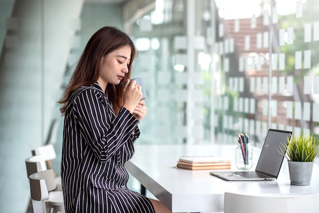 Happy young asian woman drinking coffee at table with laptop in cafe.