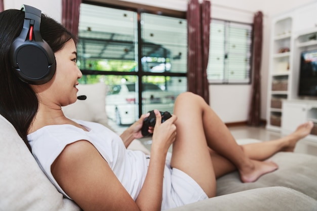 Happy young asian woman gamer with headphones playing video game and relaxing on sofa at home