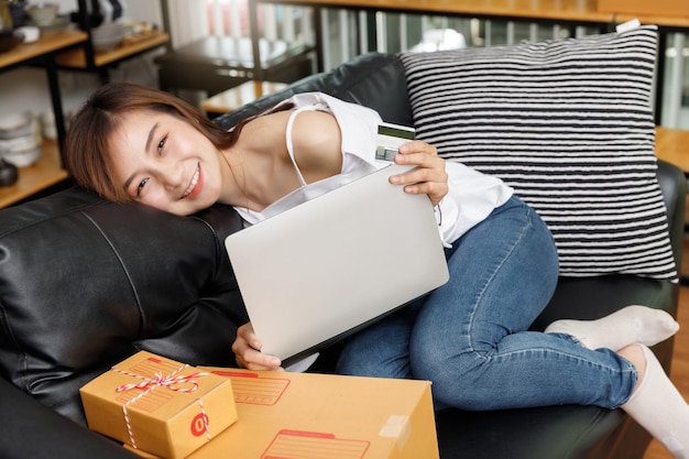 Happy young asian woman holding credit card and laptop sitting on sofa at home