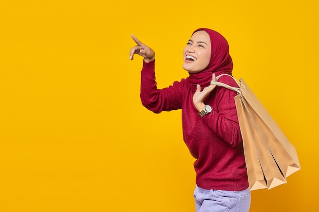 Happy young Asian woman holding shopping bag and pointing to empty space on yellow background