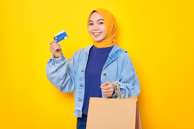 Happy young Asian woman in jeans jacket holding shopping bags and credit card looking at camera isolated over yellow background