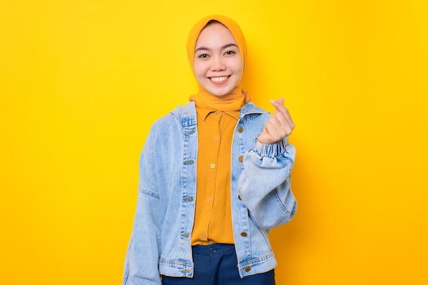 Happy young Asian woman in jeans jacket showing Korean heart gesture with two fingers crossed isolated over yellow background