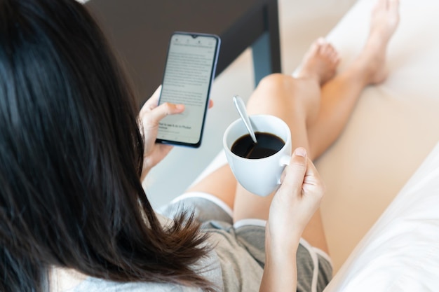 Happy young Asian woman relaxing on sofa at home in the morning while have some coffee and using smartphone Technology and lifestyle concept Top view copy spacex9