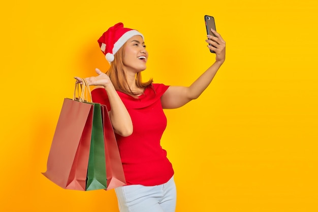 Happy young Asian woman in Santa Claus hat holding shopping bags and taking selfie on mobile