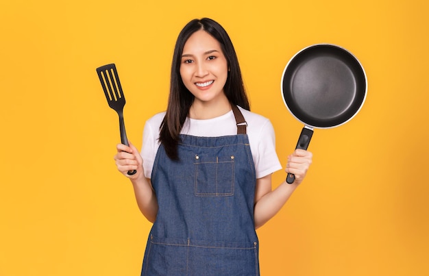 Happy young asian woman wear on an apron and hands showing spatula with pan on light yellow background.