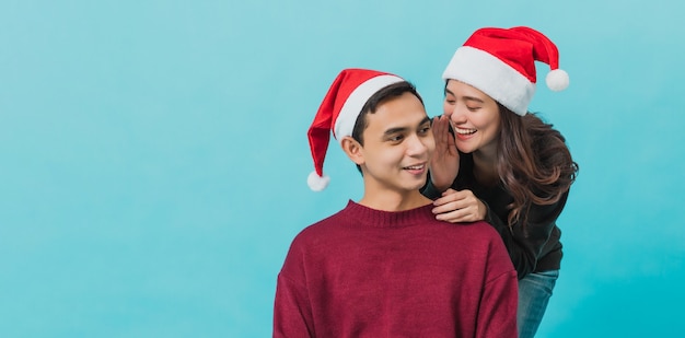 Happy young Asian woman wearing Santa Claus and whispering to her boyfriend